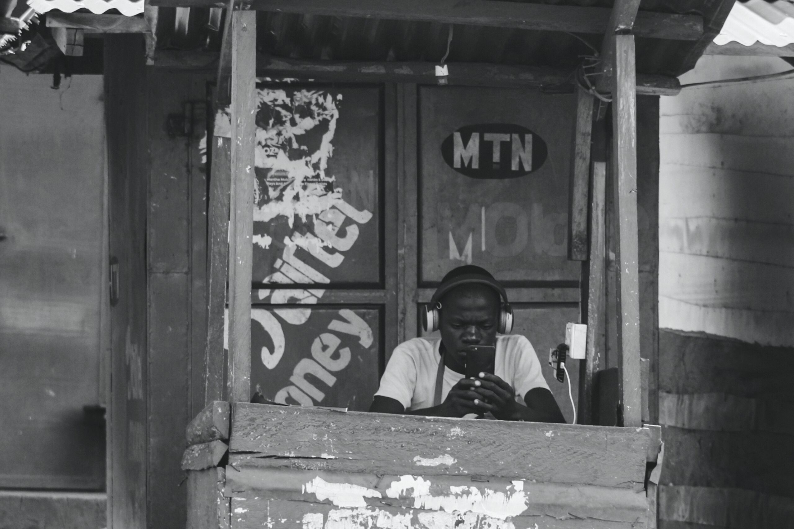 A man in a rustic airtime kiosk looks down at his phone, wearing large headphones. Greyscale, high contrast. Image by Aidah Namukose / ALT Advisory (CC BY-NC-ND).